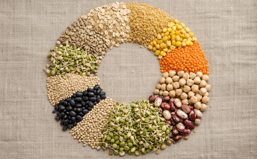 Circle of different legumes and grains arranged on beige fabric.